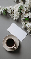 A light beige coffee cup and saucer, a blank white card, and delicate white blossoms rest on a muted gray surface