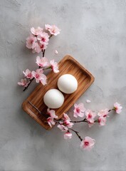 Two white spheres on a light brown wooden tray, surrounded by delicate pink cherry blossoms on a mottled gray surface