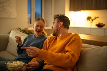 Couple laughing to tears while watching movie and joking on sofa during Friday evening at home. Man...