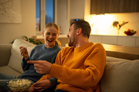 Couple laughing to tears while watching movie and joking on sofa during Friday evening at home. Man and woman curled on couch with teary eyes and wide smiles exchanging banter during film night