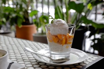 A glass of mango sticky rice topped with coconut ice cream and mint leaf, served on a white plate with a spoon, showcasing a refreshing tropical dessert.