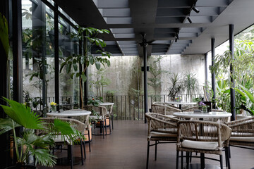 A cozy modern café with wicker chairs, round tables, lush green plants, and large glass windows letting in natural light. A geometric ceiling adds a stylish touch.