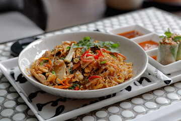 A white bowl of stir-fried glass noodles with vegetables and herbs, served alongside fresh spring rolls and dipping sauces on a patterned table.