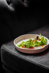 Casual salad with greens, cherry tomatoes, nuts, and croutons on a white ceramic plate, placed on a dark leather couch in a relaxed, informal dining setting