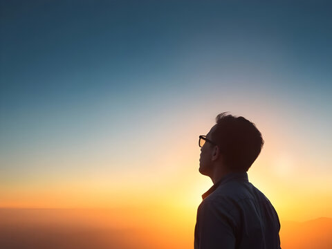 Thoughtful man in glasses looking up at a vibrant golden hour sunset or sunrise sky, contemplating future and freedom outdoors.