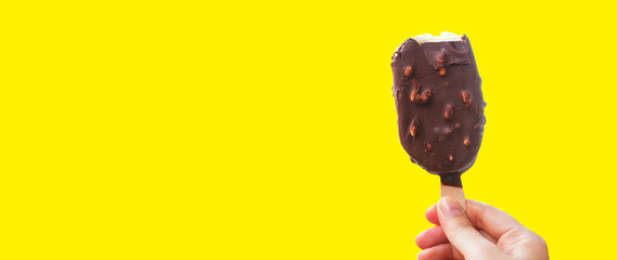 Chocolate covered ice cream bar on Yellow background, during the holidays. Hand holds delicious chocolate and nuts almond ice cream bar on a stick.