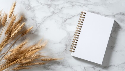 A blank notebook rests on a marble surface, beside dried wheat stalks