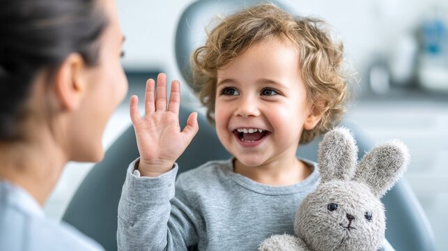 A happy toddler waves with a smile, holding a stuffed animal, while sitting in a dental chair during a checkup.