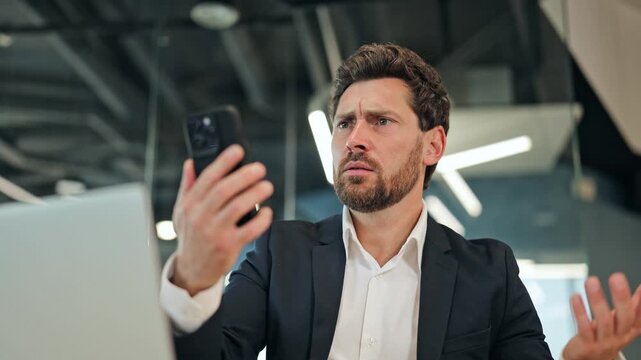 Frustrated professional businessman, dressed in a suit, reacts to smartphone content in a modern office. Displaying clear confusion annoyance, the male struggles with technology, feeling overwhelmed.