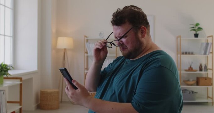 Fat overweight man with glasses holds smartphone, squinting at screen as he tries reading, struggling for focus and better vision. Man with farsightedness eye disease squints at phone screen
