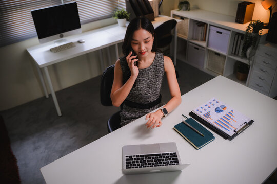 Young businesswoman talking on smartphone at office desk, checking time on smartwatch, analyzing business data on laptop