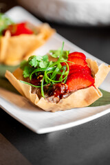 Gourmet appetizer in crispy shell with savory filling, garnished with cilantro, green onion, and strawberry slices. Elegant plating on white dish, fusion cuisine concept