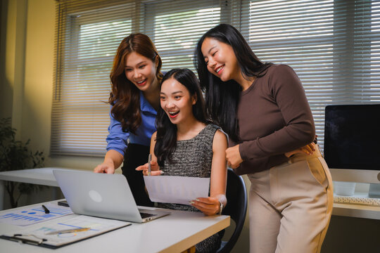 Asian women colleagues collaborating on project, looking at laptop, celebrating business success in modern office