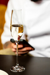 Close-up of a champagne flute with sparkling wine on a dark surface, blurred background with person holding smartphone, elegant moment of leisure or celebration.
