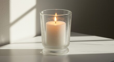 A soft, glowing white candle sits in a frosted glass votive, casting gentle shadows on a white surface.