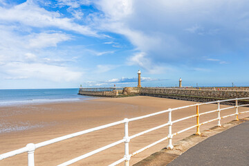 A pier with lighthouses stretches out to sea with a beach and railings in the foreground.