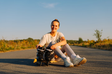 Fototapeta premium A young woman sits on the ground, smiling and resting her inline skates by a scenic road under a clear blue sky at sunset