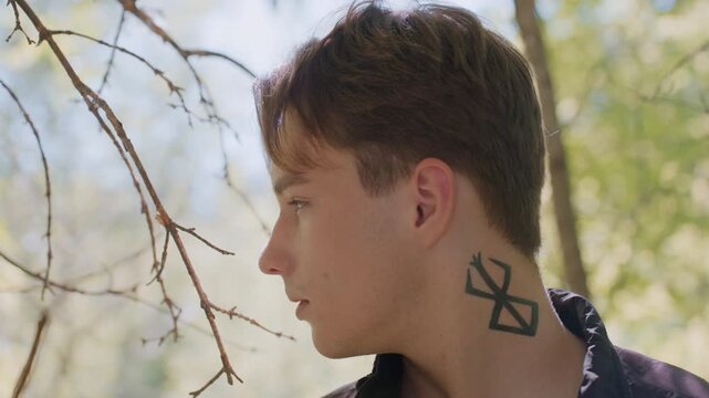 Warrior stands quietly in forest, making silent observation by shifting eyes left and right without turning head, focused and cautious as sunlight filters through trees highlighting tattoo on neck