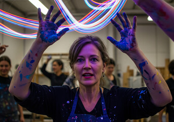 A woman with paint-covered hands raises them in the air, with light trails swirling above her, surrounded by other people in a studio setting.