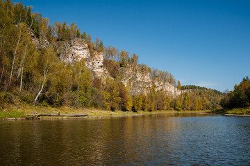 A beautiful lake, perfectly nestled amidst lush green trees and rugged rocks, shines brightly under the warmth of the sun on this clear day, creating a serene, tranquil scene