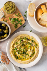 Creamy hummus is beautifully plated with green herbs and olive oil, surrounded by fresh olives, pita bread, and various seasonings on a simple table