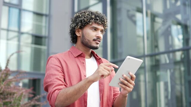 Young confident businessman uses digital tablet while standing on the street near office building. Handsome male in red shirt chats online with client, browses web, writes or reads messages. Close up
