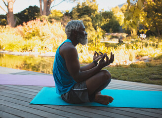 Senior African American man practicing meditation on turquoise yoga mat on wooden deck beside pond