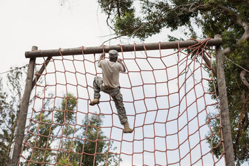 African American male soldier in camo climbing red rope net reaching wooden beam on obstacle course