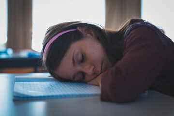 Female child resting head on open lined notebook at desk in classroom wearing pink headband