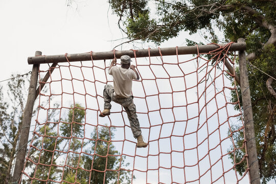 African American male soldier in camo climbing red rope net reaching wooden beam on obstacle course