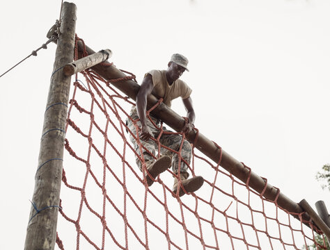 African American male soldier climbing red rope net obstacle course gripping beam and hoisting legs