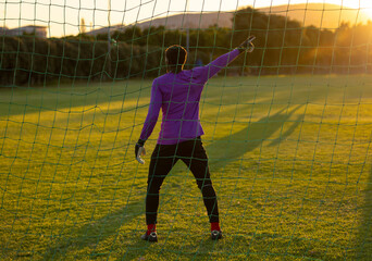 Teenage male goalkeeper wearing padded gloves, pointing on grass pitch by soccer net at golden hour