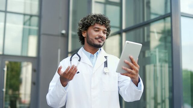 Young smiling doctor talking online via video call using digital tablet while standing outside near hospital building. Medical worker physician in white coat communicates remotely, consults patient