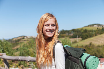 Young woman smiling, carrying a backpack and sleeping mat, looking away with hills in background