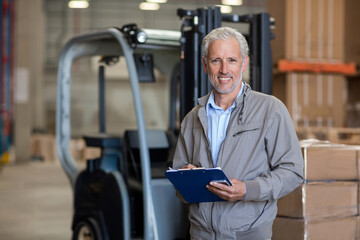 Senior male worker standing beside forklift and holding clipboard near shelving racks in warehouse