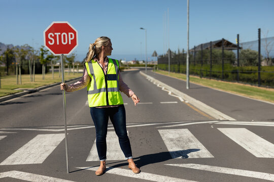 Female crossing guard holding stop sign directing traffic on roadside crosswalk wearing safety vest - Powered by Adobe