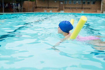Child swimmer wearing blue cap, pink swimsuit floating on yellow noodles in pool with lifebuoys