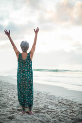 Barefoot senior woman raising arms toward sky on sandy beach by calm sea, under cloudy sky