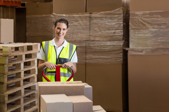 Warehouse worker operating pallet jack loaded with boxes among pallet stacks in warehouse - Powered by Adobe