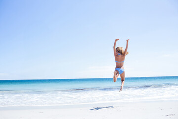 Female beachgoer jumping mid-air on sandy beach under clear blue sky with turquoise ocean waves