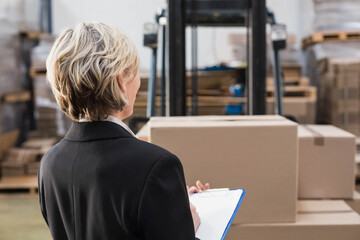 Senior woman standing with back facing viewer in warehouse wearing blazer holding clipboard and pen