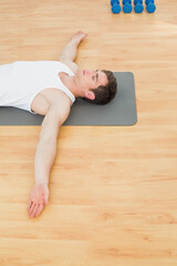 Fototapeta premium Man wearing white tank top and shorts lying on exercise mat in gym near blue dumbbells