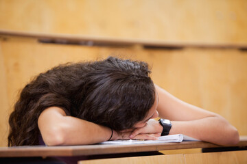 female student resting head on arms atop open notebook with digital wristwatch on desk in classroom