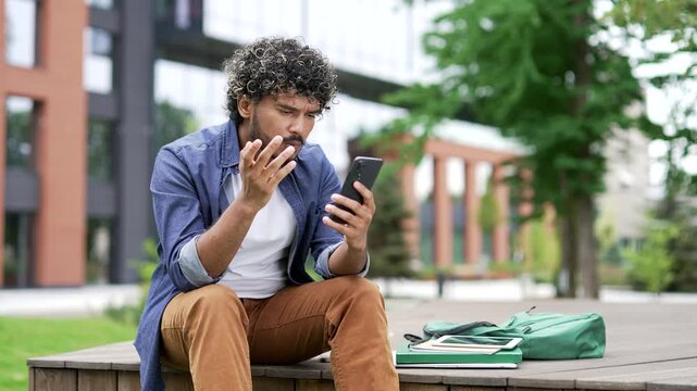 Worried male student read bad news on mobile phone while sitting on a bench in campus space near university building. Frustrated stressed handsome man upset looking at message on smartphone. Close up