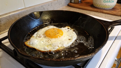 Golden yolk fried egg with bubbling whites in skillet, rustic stovetop kitchen breakfast preparation photography.