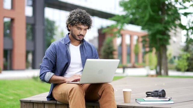Male student works on a laptop while sitting on a bench in campus space near the university building. Young man studies using a computer. Handsome freelancer is chatting online, communicating remotely