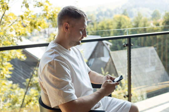 Casual man sitting on balcony in white clothes and looking at smartphone with scenic view of nature and houses in background. Concept of modern lifestyle, technology, and relaxation. Symbol of digital
