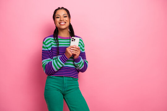 Confident young woman with braided hair holding smartphone against bright pink background, dressed in colorful striped sweater - Powered by Adobe