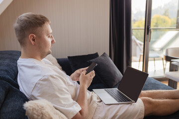 Young man sitting on sofa with laptop and smartphone, working remotely or managing online tasks. Concept of freelancing, digital lifestyle, mobile technology, e-banking, online shopping, communication