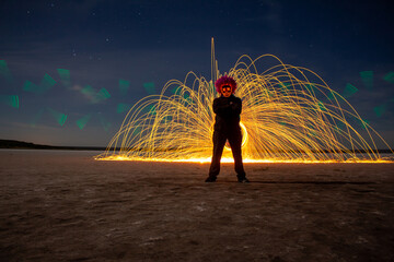Person wearing glowing mask with arms crossed standing before sparks of light at night.